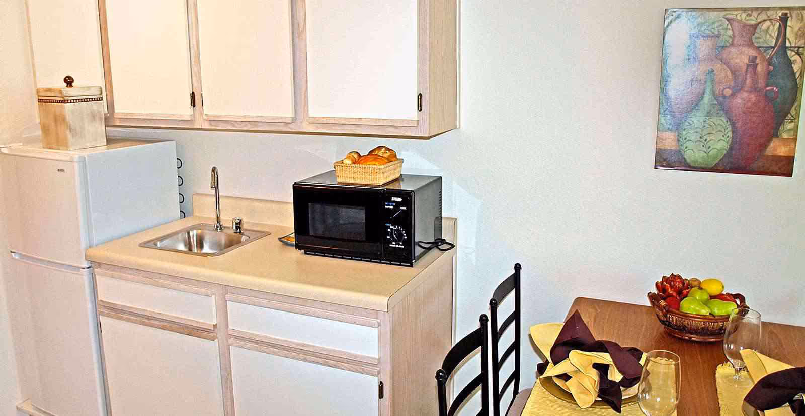 Small kitchen area with a white refrigerator, a countertop with a sink, a black microwave, and a basket of bread on top of the microwave. Adjacent to the kitchen is a dining table set with folded napkins, glasses, and a fruit basket. A colorful painting of vases hangs on the wall above the dining table.