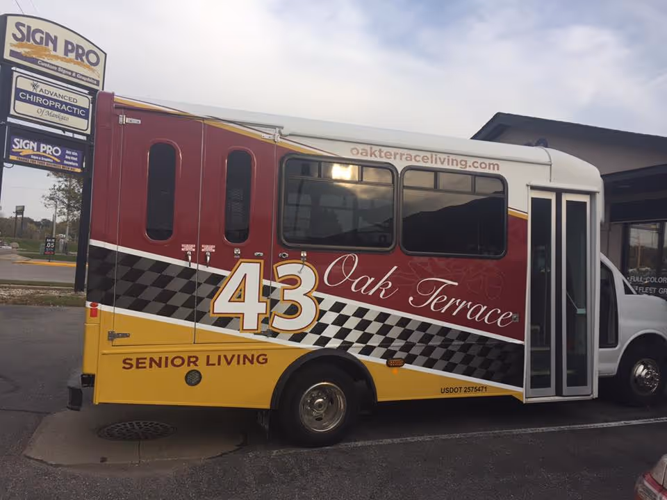 Side view of a small shuttle bus with maroon, yellow, and white colors, displaying the number 43 and the text Oak Terrace Senior Living. The bus is parked in a lot near a sign for Sign Pro and Advanced Chiropractic.