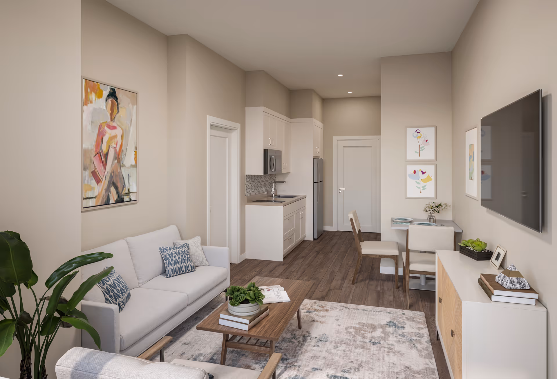 A modern living room and kitchenette area with a light gray sofa, wooden coffee table with books and a plant, a small dining table with two chairs, a wall-mounted flat screen TV, and light-colored walls decorated with abstract and floral artwork. The floor is wooden and there is a large plant in the corner.