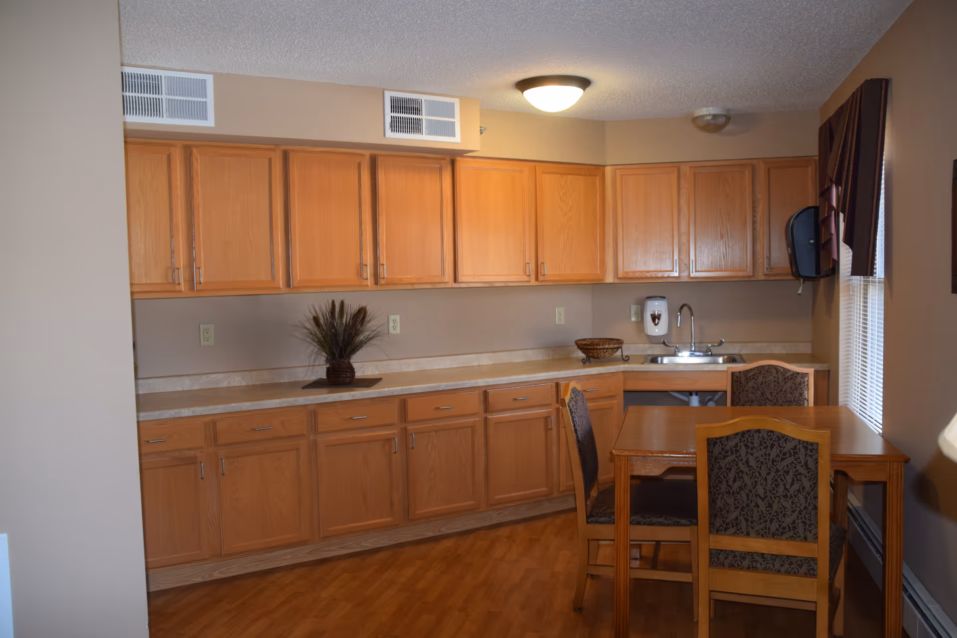 A kitchen area with wooden cabinets along the walls, a countertop with a small plant and a basket, a sink with a soap dispenser, and a wooden dining table with four chairs. There is a window with blinds and a valance on the right side.