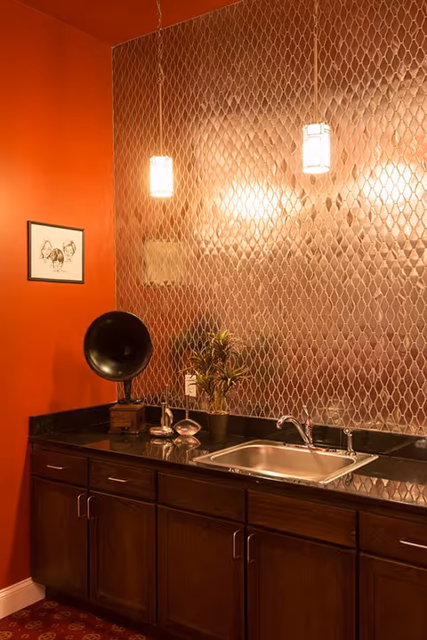 A kitchen area with dark wooden cabinets, a stainless steel sink, and a black countertop. The backsplash features a textured, diamond-patterned metallic surface. Two pendant lights hang from the ceiling, illuminating the space. On the countertop, there is a vintage gramophone, a small plant, and decorative items. The wall to the left is painted a warm orange color and has a framed black and white drawing of three faces.