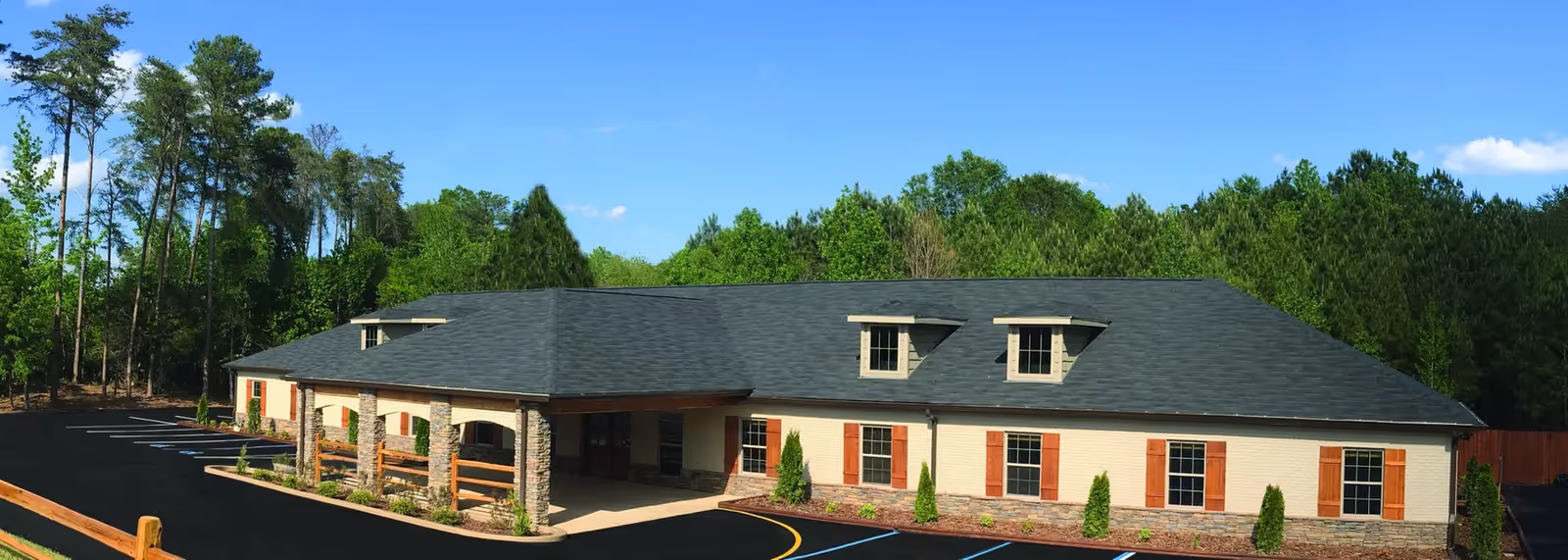Single-story assisted living building with a covered entrance, parking lot, and trees under a blue sky.