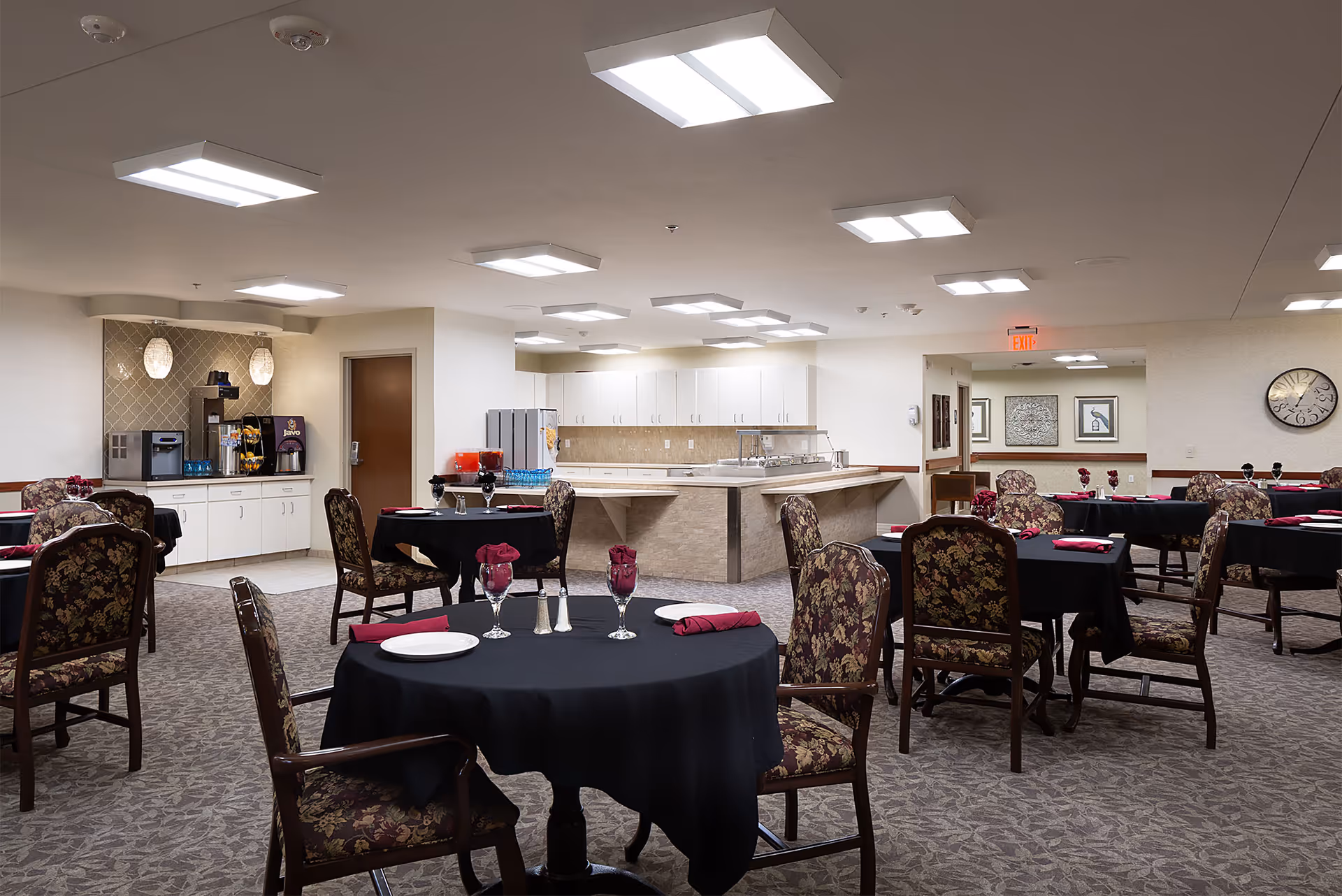 A spacious dining room with multiple round tables covered with black tablecloths, each set with plates, glasses, and folded red napkins. The chairs have floral upholstery. In the background, there is a counter area with a beverage station including a coffee machine and juice dispensers. The room is well-lit with ceiling lights and has a carpeted floor.