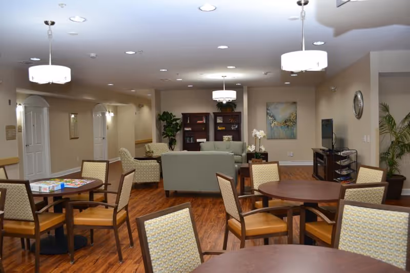 Communal senior living activity room with round tables and chairs in the foreground and a seating area with sofas, bookshelf, and TV in the background.
