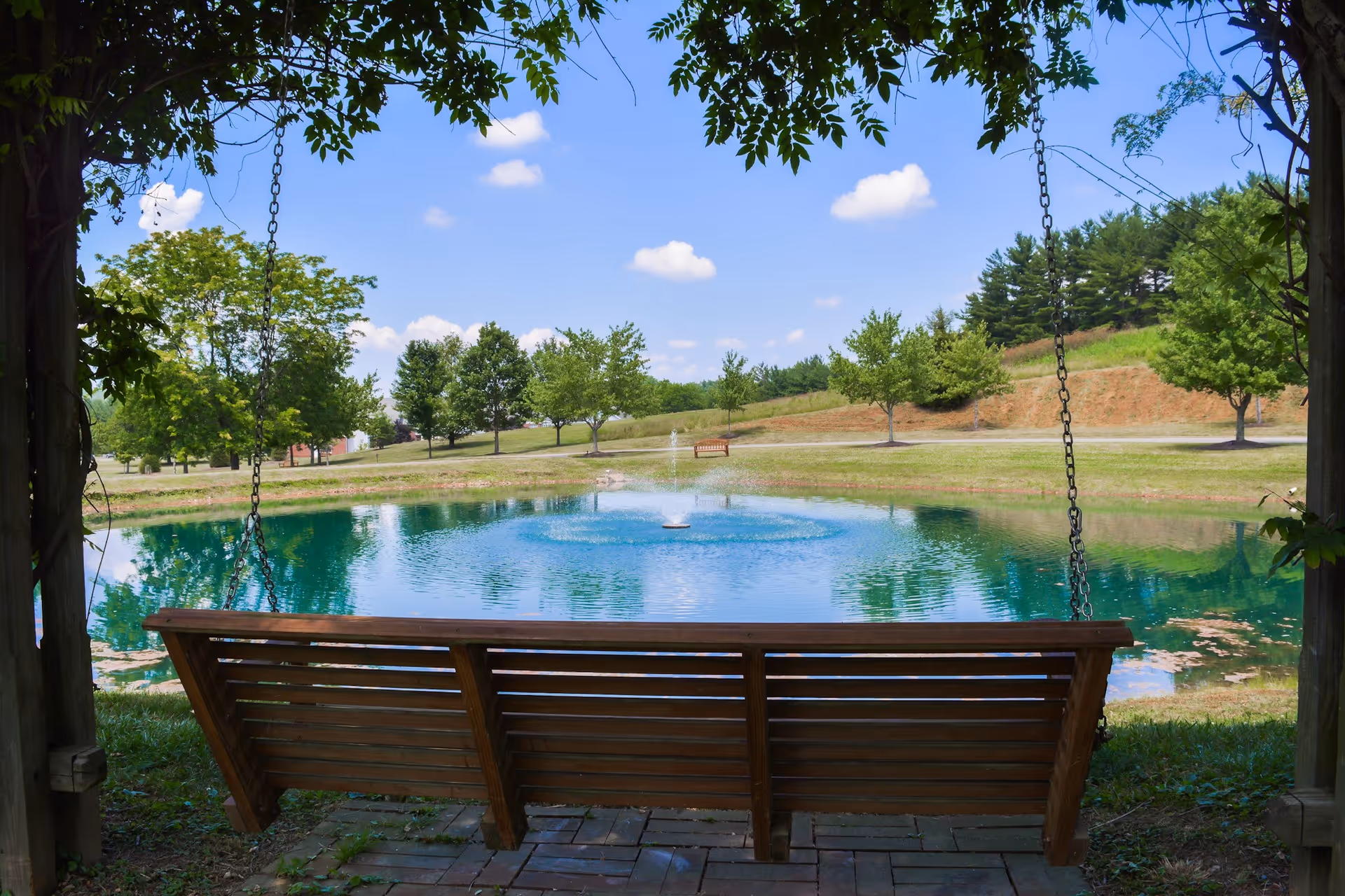 A wooden swing bench under a shaded pergola overlooking a small pond with a fountain and trees on a sunny day.