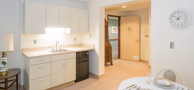 Small kitchenette with white cabinets, sink, and mini-fridge next to a dining table and an open doorway to a hallway.
