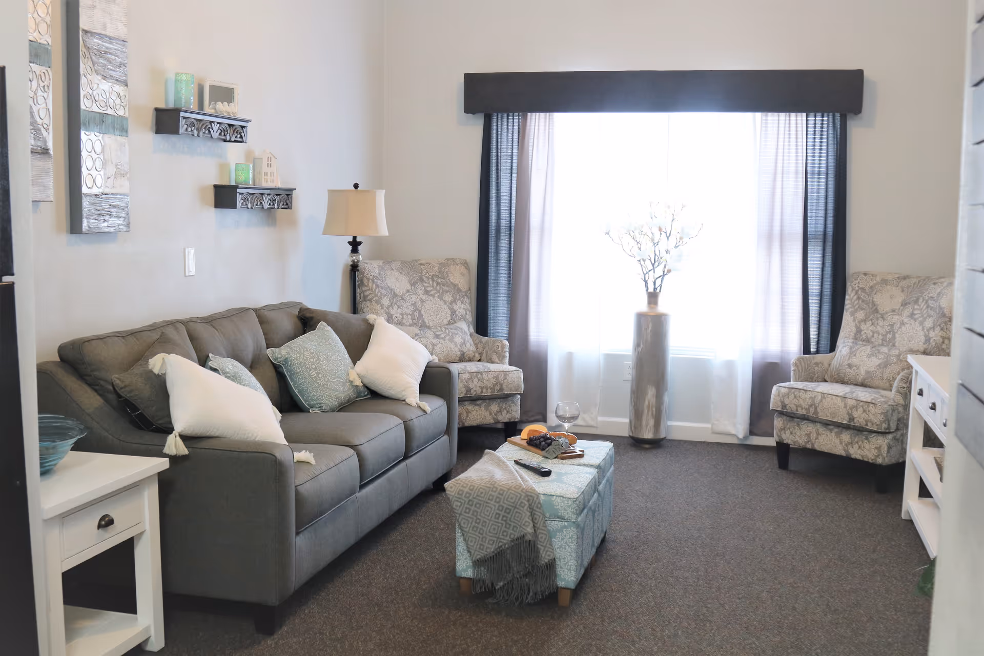 A cozy living room with a gray sofa adorned with white and blue pillows, two patterned armchairs, a small ottoman with a throw blanket, a side table with a lamp, and a large window with sheer curtains letting in natural light. Decorative shelves and artwork are on the wall.