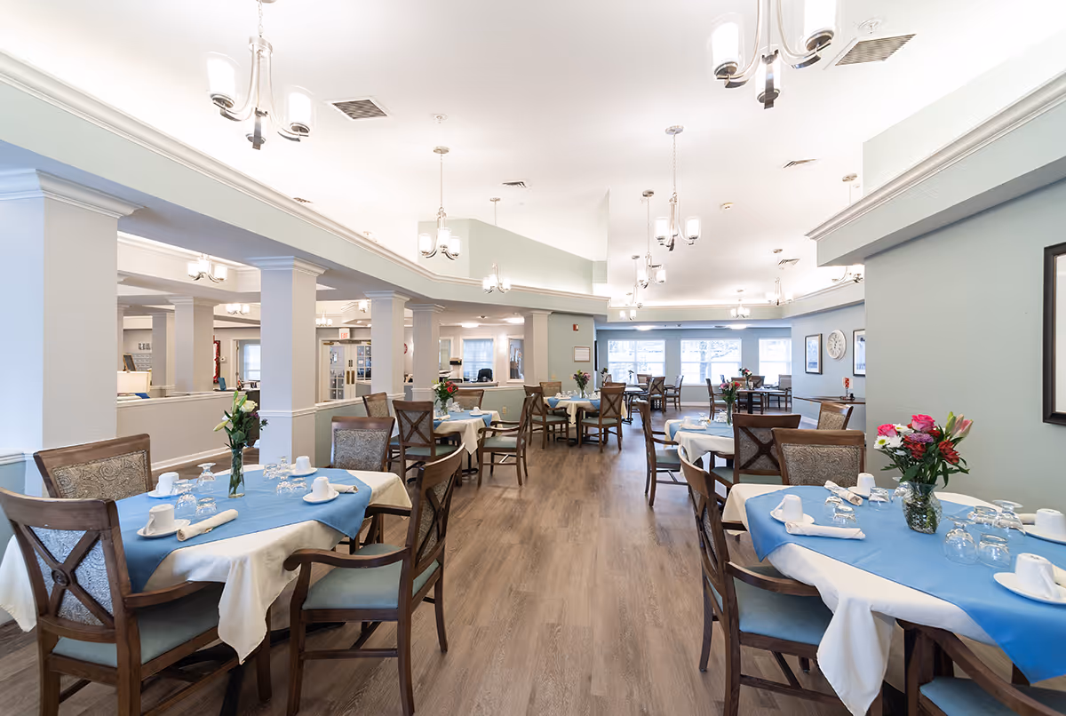 A bright and spacious dining room with multiple tables covered in white and blue tablecloths, each set with cups, glasses, and napkins. Wooden chairs with cushioned seats surround the tables. The room features wooden flooring, light blue walls, and several ceiling light fixtures. Vases with fresh flowers are placed on the tables, and large windows at the far end allow natural light to fill the space.