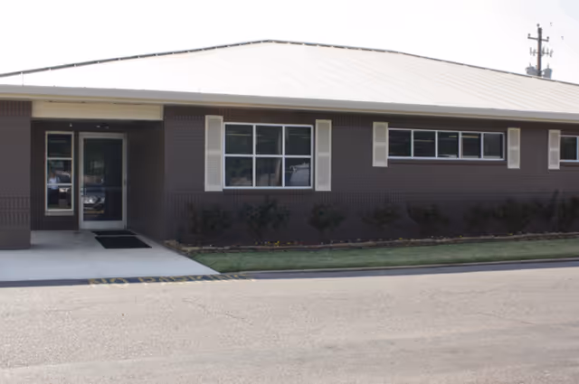 Exterior view of a single-story building with a metal roof, brown brick walls, white window shutters, and a glass door entrance. There is a small garden with shrubs in front of the building and a paved driveway with 'NO PARKING' painted on it.
