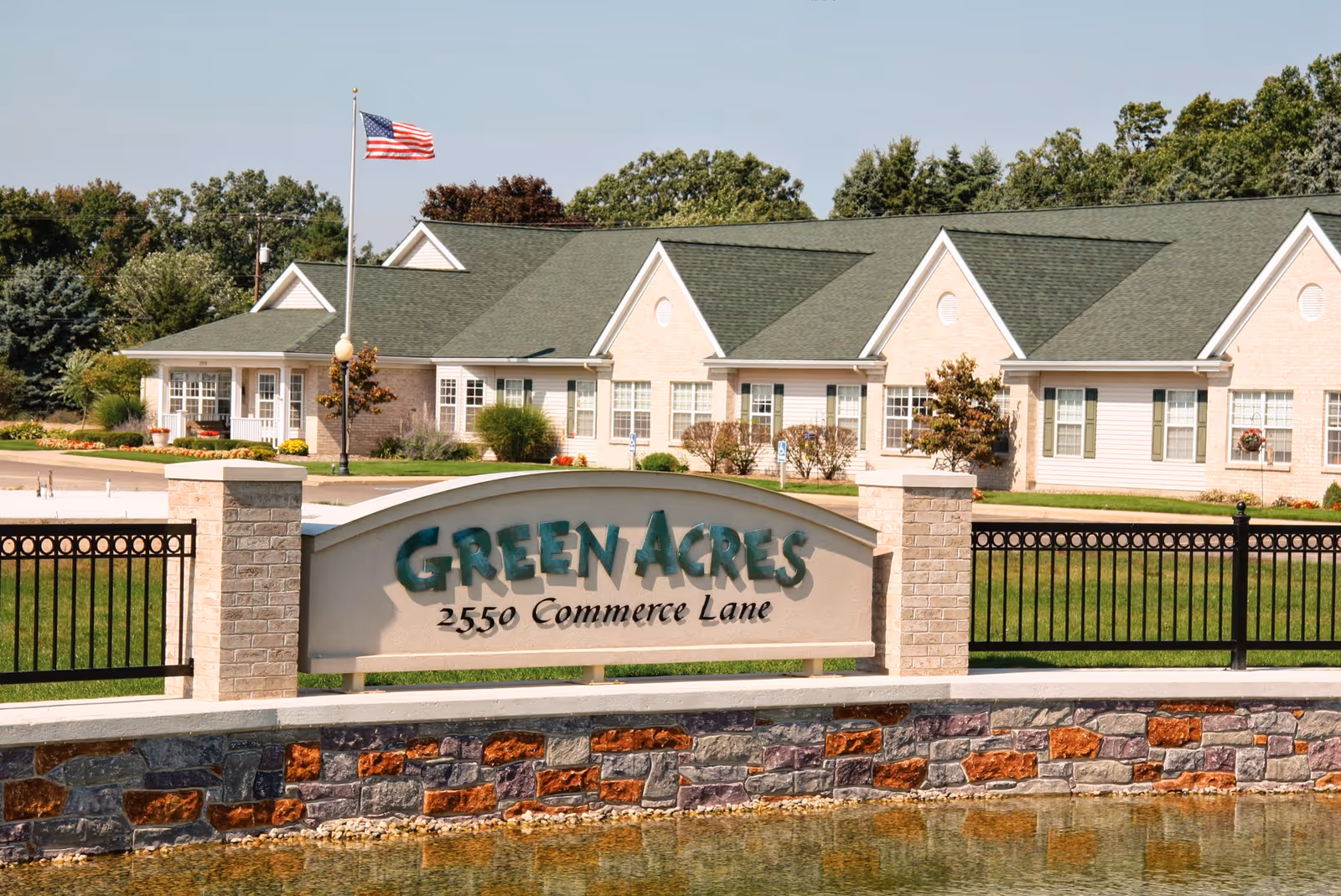 Exterior view of Green Acres Ionia facility showing a single-story building with green roofs and beige walls, an American flag on a flagpole, a landscaped lawn, and a stone and brick sign in front that reads 'Green Acres 2550 Commerce Lane'.
