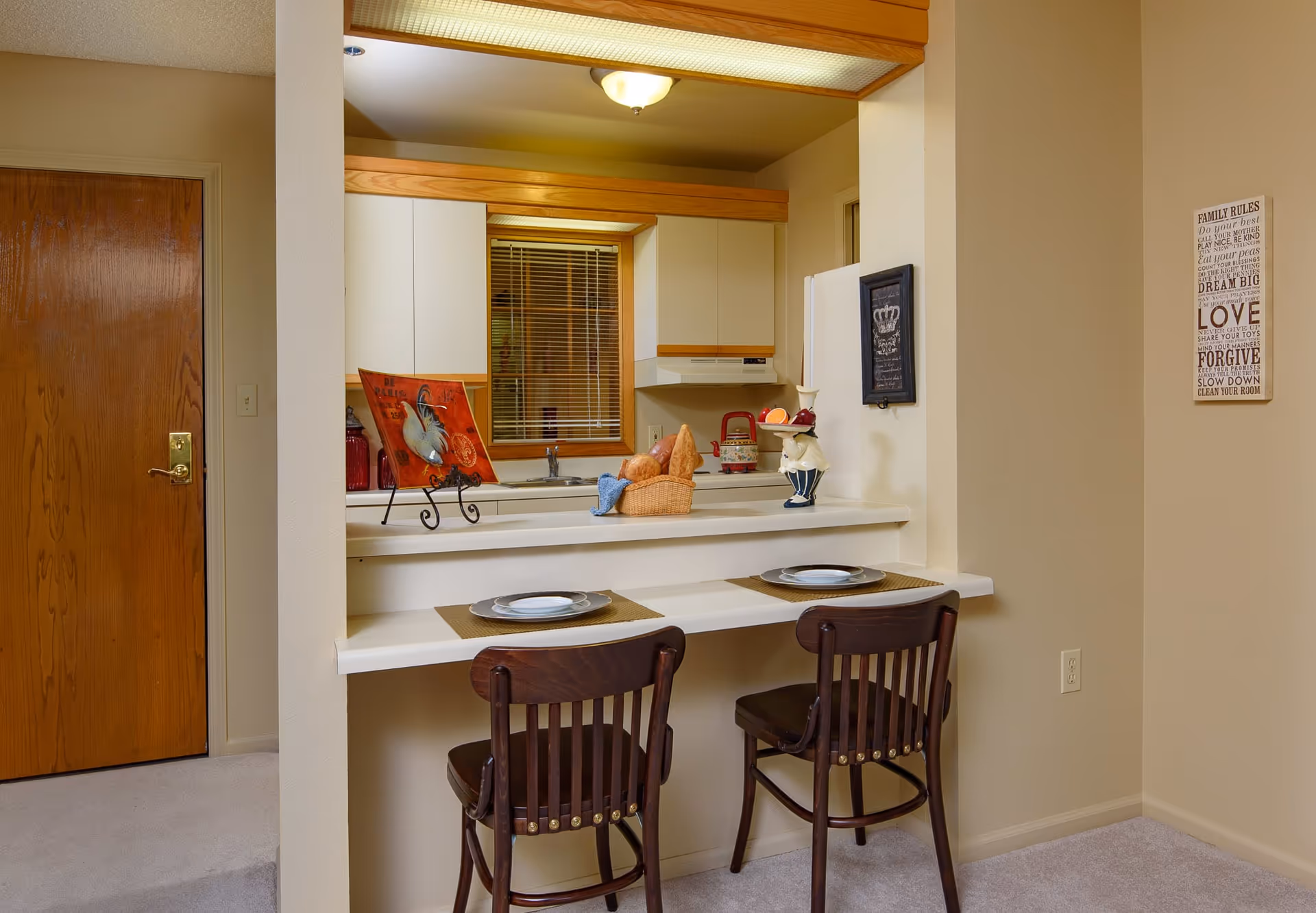 Interior view of a small kitchen area with a breakfast bar featuring two dark wooden chairs and place settings. The kitchen has white cabinets, a window with blinds, and various decorative items including a basket with bread and a ceramic chef figurine. Beige walls and carpeted floor surround the space.