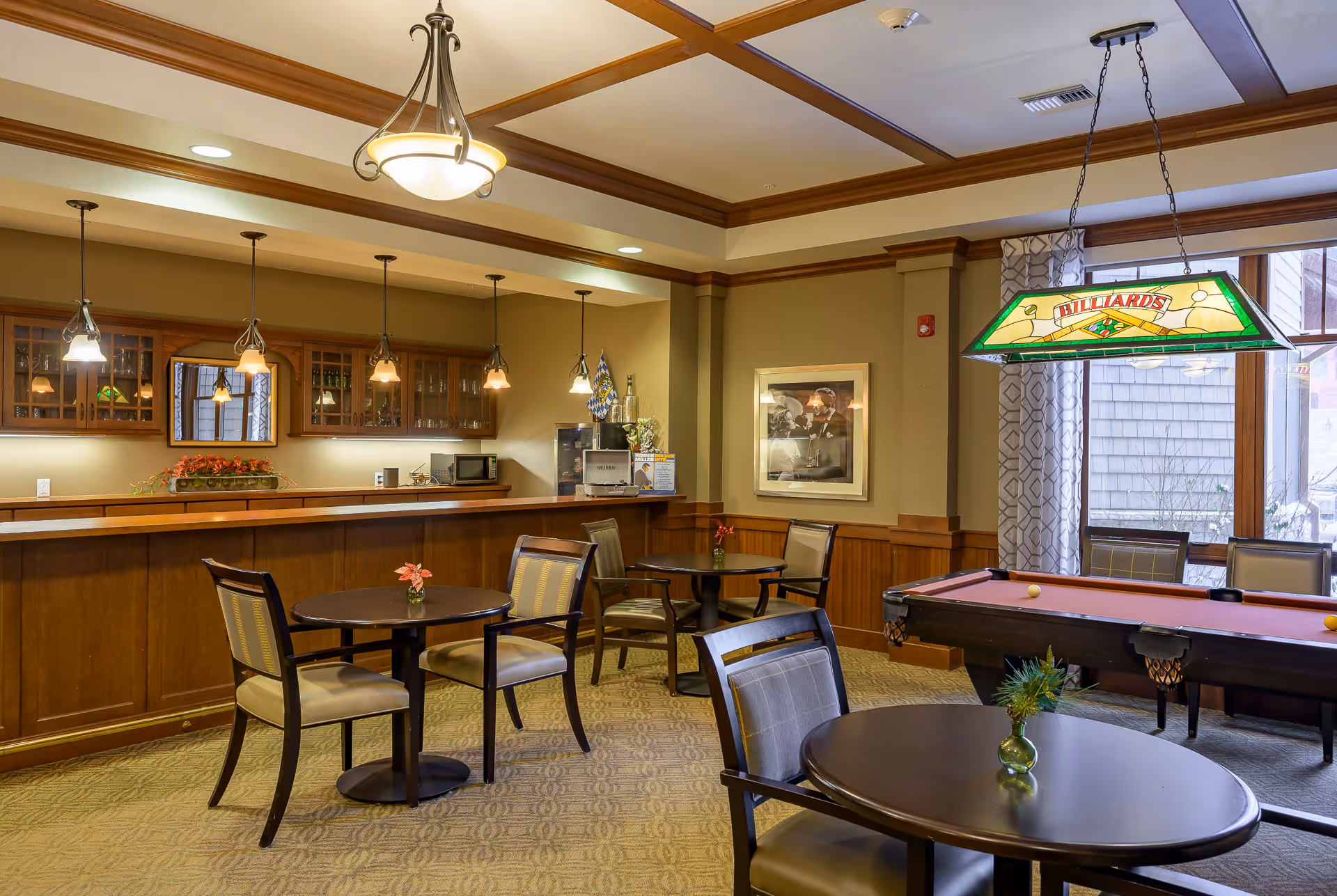 A cozy common area in an assisted living facility featuring a pool table with a stained glass 'Billiards' light hanging above it, several round tables with chairs, a wooden bar with hanging pendant lights, and a window with patterned curtains letting in natural light.