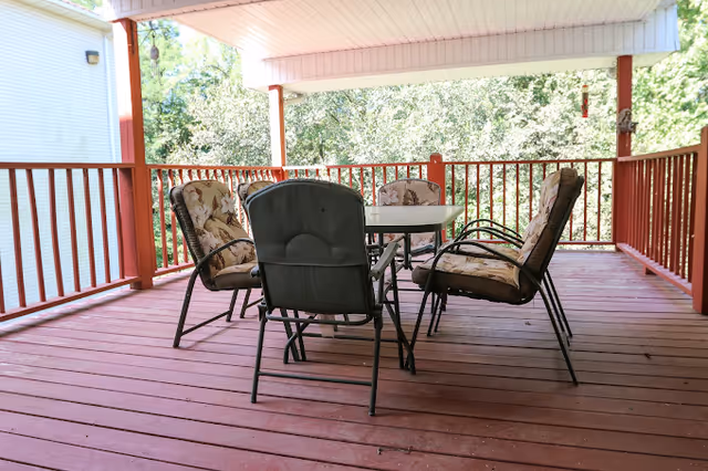 Covered red wooden deck with a table and six cushioned patio chairs overlooking trees.
