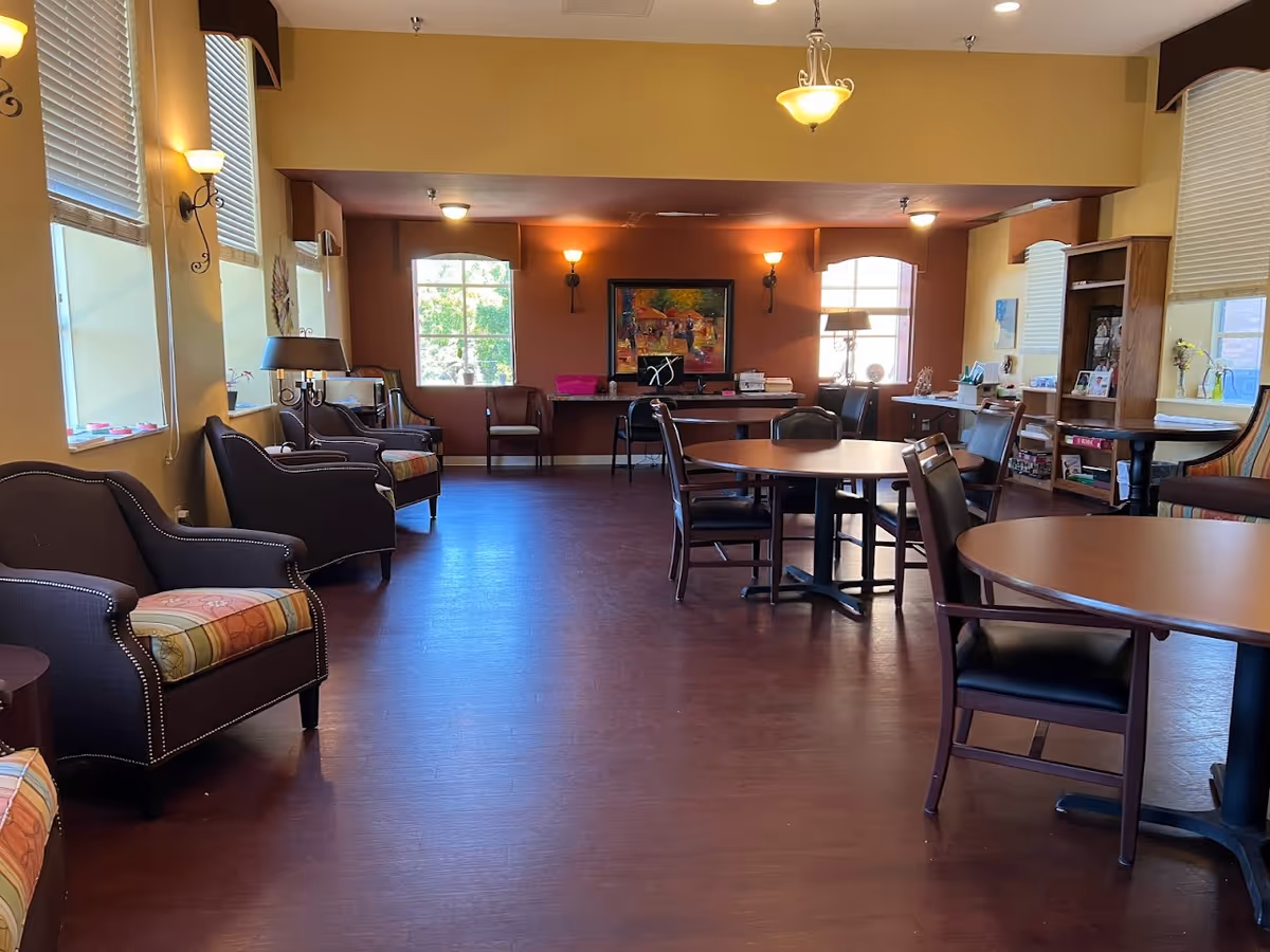 A spacious senior living common room with armchairs, round tables, bookshelves and large windows under warm lighting.