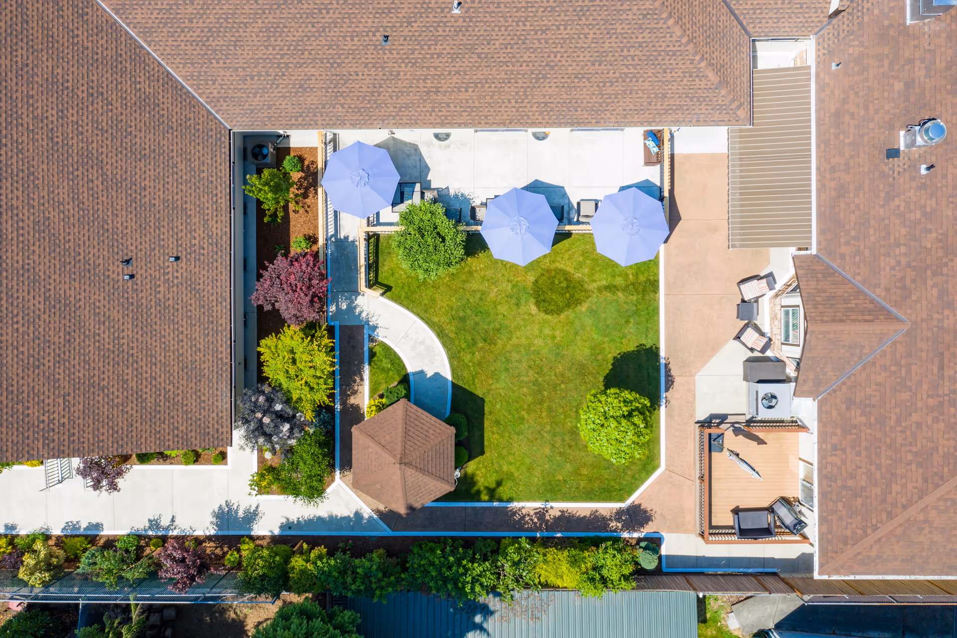 Aerial view of a senior living facility courtyard with a green lawn, three blue patio umbrellas, a small gazebo, various trees and shrubs, and surrounding buildings with brown roofs.