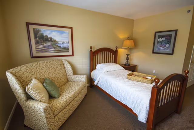 A neatly furnished assisted living bedroom with a single wooden bed, nightstand and lamp, and a patterned loveseat under framed artwork.
