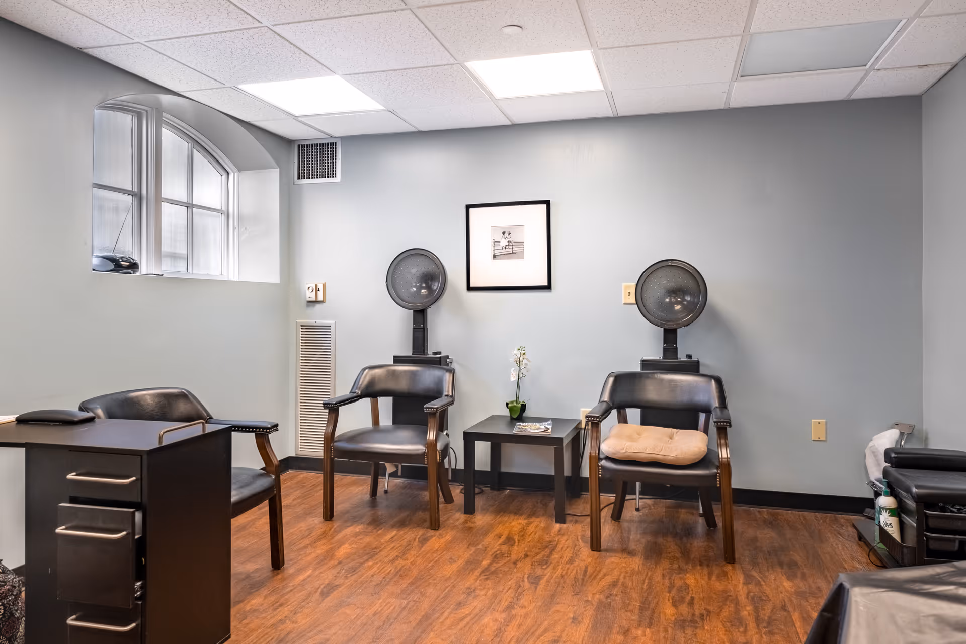 A small room with two black salon chairs positioned under hair dryers, a small black table with a potted plant and magazine between them, a black desk with a chair to the left, and a window with an arched top on the left wall. The room has light gray walls and wood flooring.