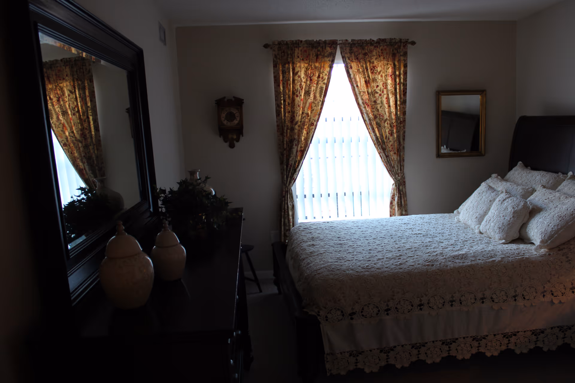 A bedroom with a bed covered in a white lace bedspread and multiple pillows. There is a window with floral curtains letting in light, a dark wooden dresser with decorative vases, a wall clock, and a mirror on the wall.