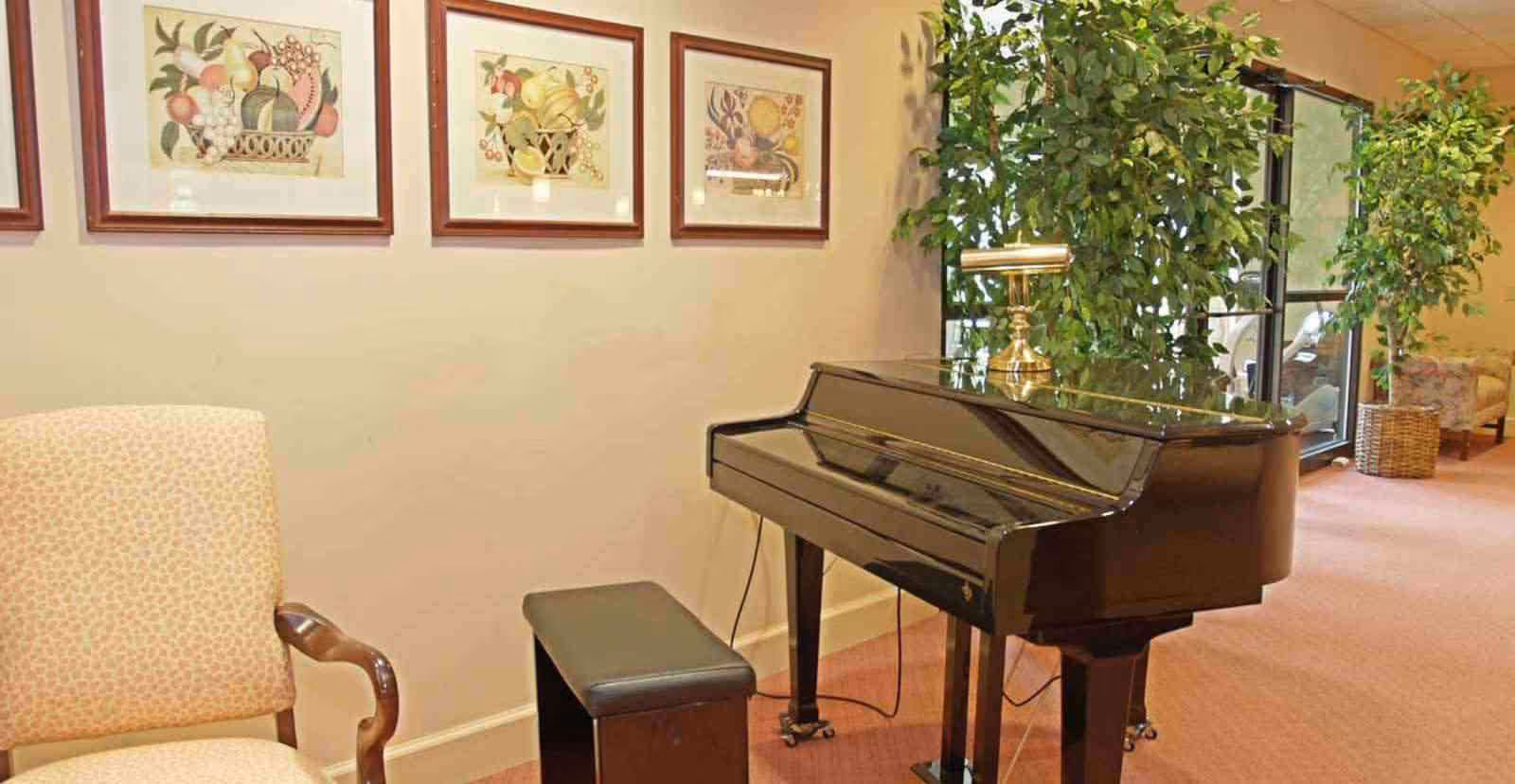 Interior view of a senior living facility lounge area featuring a black grand piano with a matching bench, a beige upholstered armchair, framed fruit artwork on the wall, and large potted plants near glass doors leading outside.