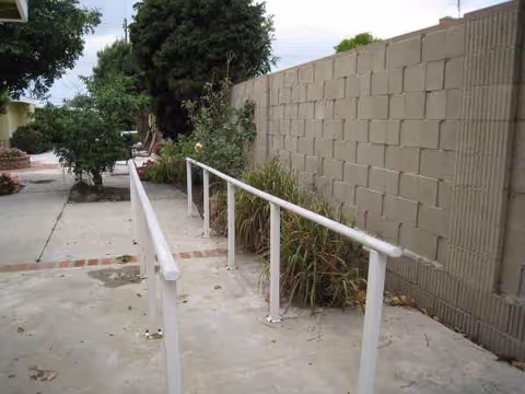 Outdoor walkway with white handrails along a concrete path beside a tall block wall and plants.