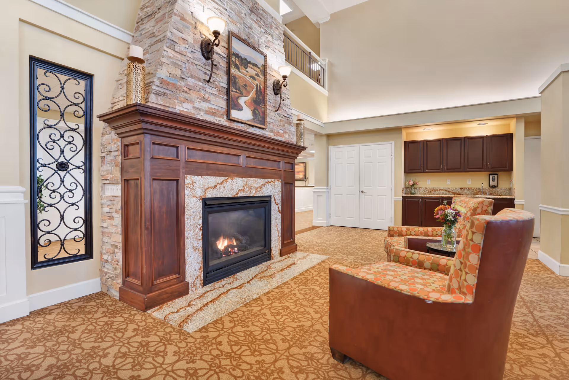 A cozy interior common area featuring a stone and wood fireplace with a lit fire, two patterned armchairs with a small round table holding a vase of flowers, beige walls, and a carpet with a decorative pattern. There is a small kitchenette with dark wood cabinets and granite countertops in the background.