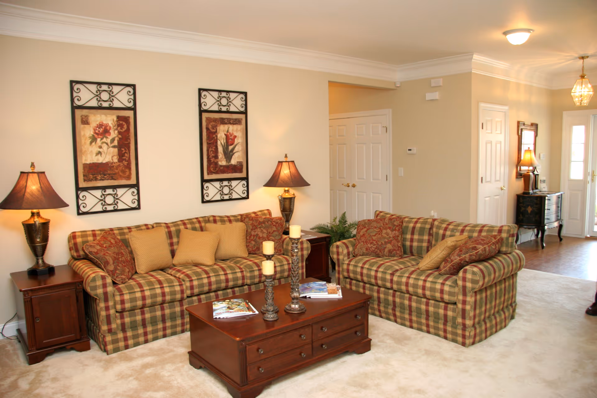 A cozy living room with two plaid sofas adorned with patterned and solid cushions. Between the sofas is a wooden coffee table with decorative candles and magazines. Two table lamps with dark shades sit on wooden side tables beside each sofa. The walls are light beige with two framed floral artworks hanging above one sofa. The room has a beige carpet and an open doorway leading to a hallway with additional furniture and lighting fixtures.