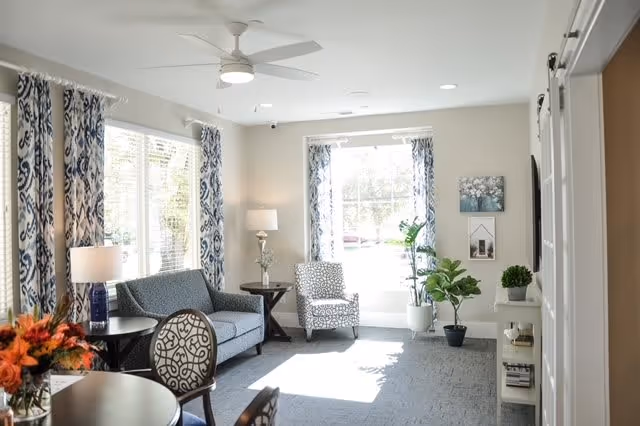 A bright and airy living room with large windows covered by patterned blue and white curtains. The room features a blue sofa, a patterned armchair, a ceiling fan with light, a side table with a lamp, several potted plants, and a small white shelving unit with decorative items. The walls are light-colored, and the floor is carpeted in gray.