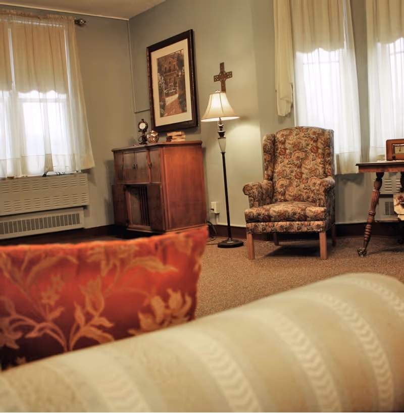 A cozy living room area with a floral patterned armchair, a wooden side table, a floor lamp, and a wooden cabinet against a light blue wall. The room has two windows with sheer white curtains, a framed picture on the wall, and a decorative cross above the lamp. In the foreground, there are parts of a beige striped sofa and a red floral pillow.