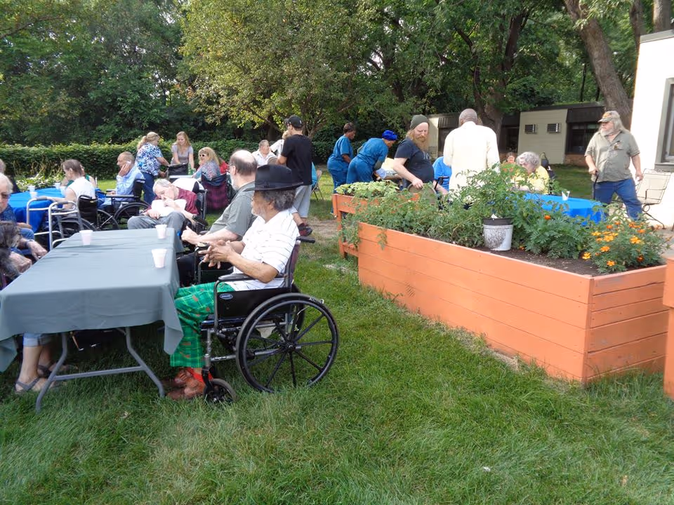 A group of elderly people and caregivers gathered outdoors in a garden area with green grass and trees. Some elderly individuals are seated at tables covered with blue and gray tablecloths, while others are in wheelchairs. Raised garden beds with plants and flowers are visible on the right side. The setting appears to be a social or recreational event at a senior living facility.