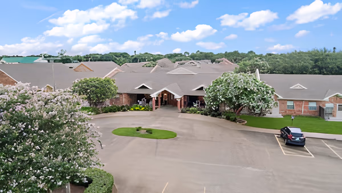 Front exterior view of Morada Victoria facility showing a large building with multiple roof peaks, surrounded by green trees and bushes, a circular driveway with a small grassy island, and a parked black car in the parking lot under a partly cloudy blue sky.