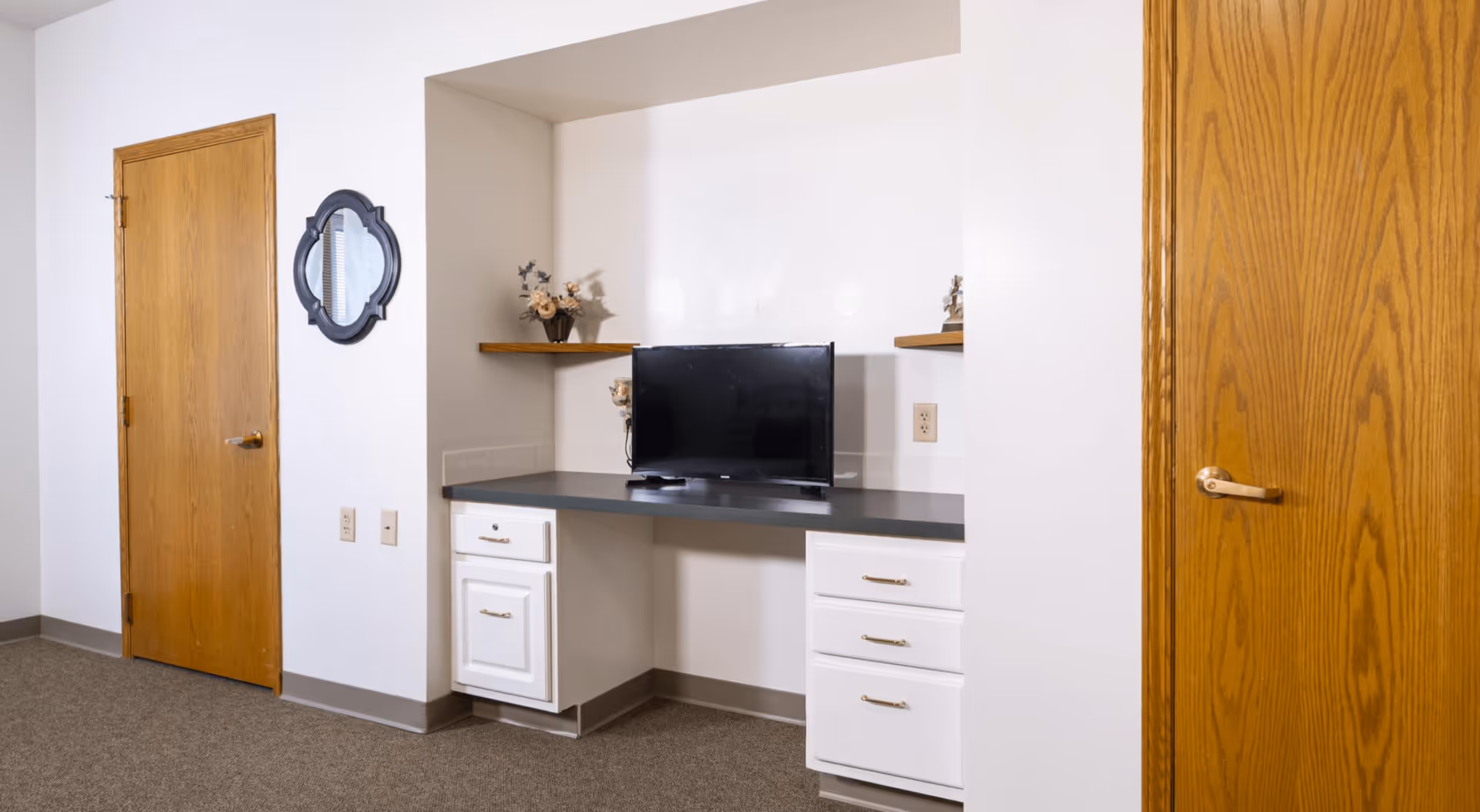 Interior view of a room with two wooden doors on either side, a black flat-screen TV placed on a built-in desk with white drawers, two wooden shelves with decorative items, a small decorative mirror on the wall, and beige carpeted floor.
