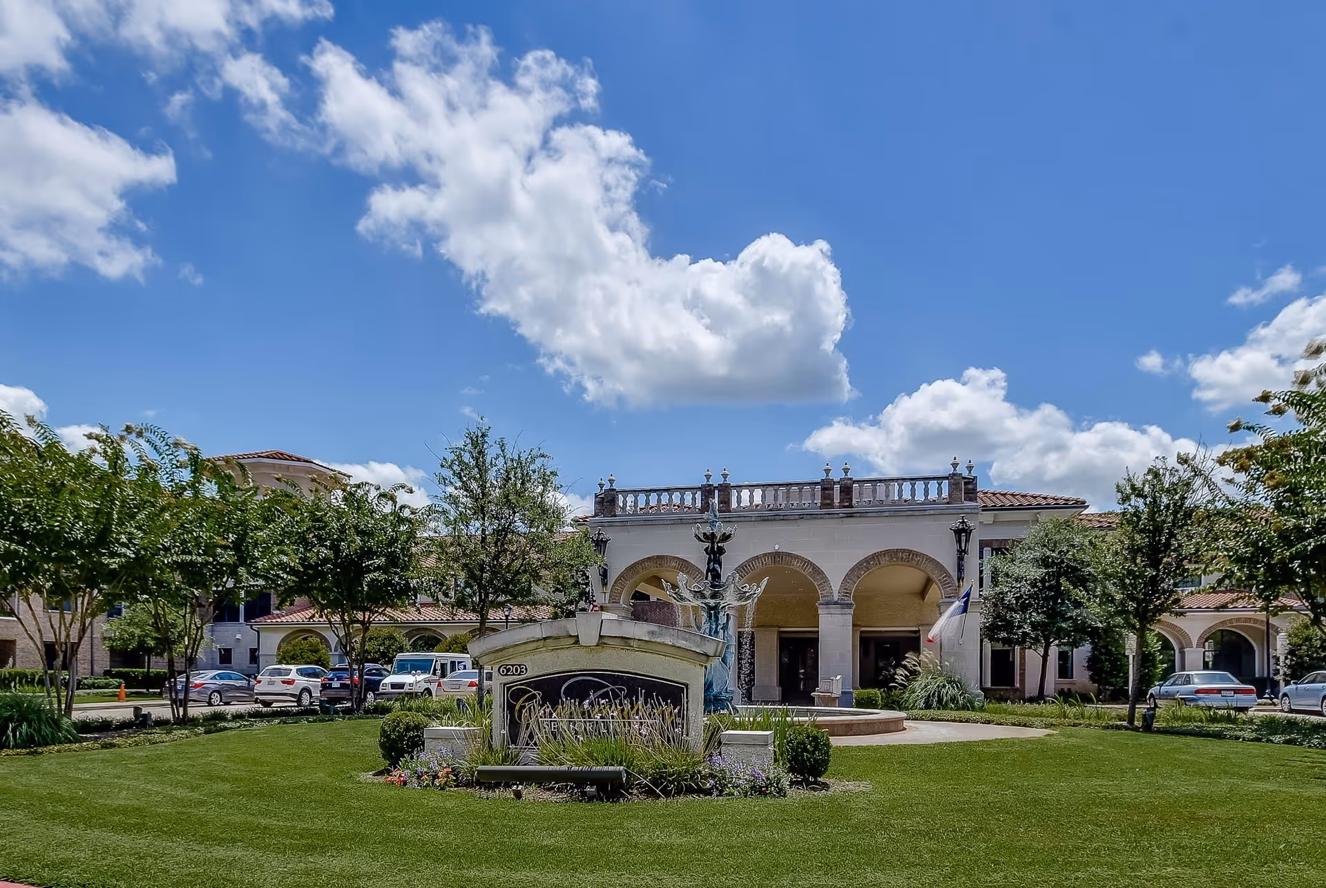 Exterior view of Conservatory At Champion Forest senior living facility showing a large building with arched entrances, a fountain in front, well-maintained green lawn, trees, and several parked cars under a partly cloudy blue sky.