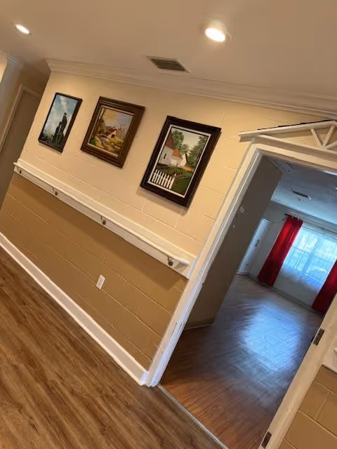 Interior hallway with framed paintings, a handrail along the wall, and an open doorway showing a room with red curtains.