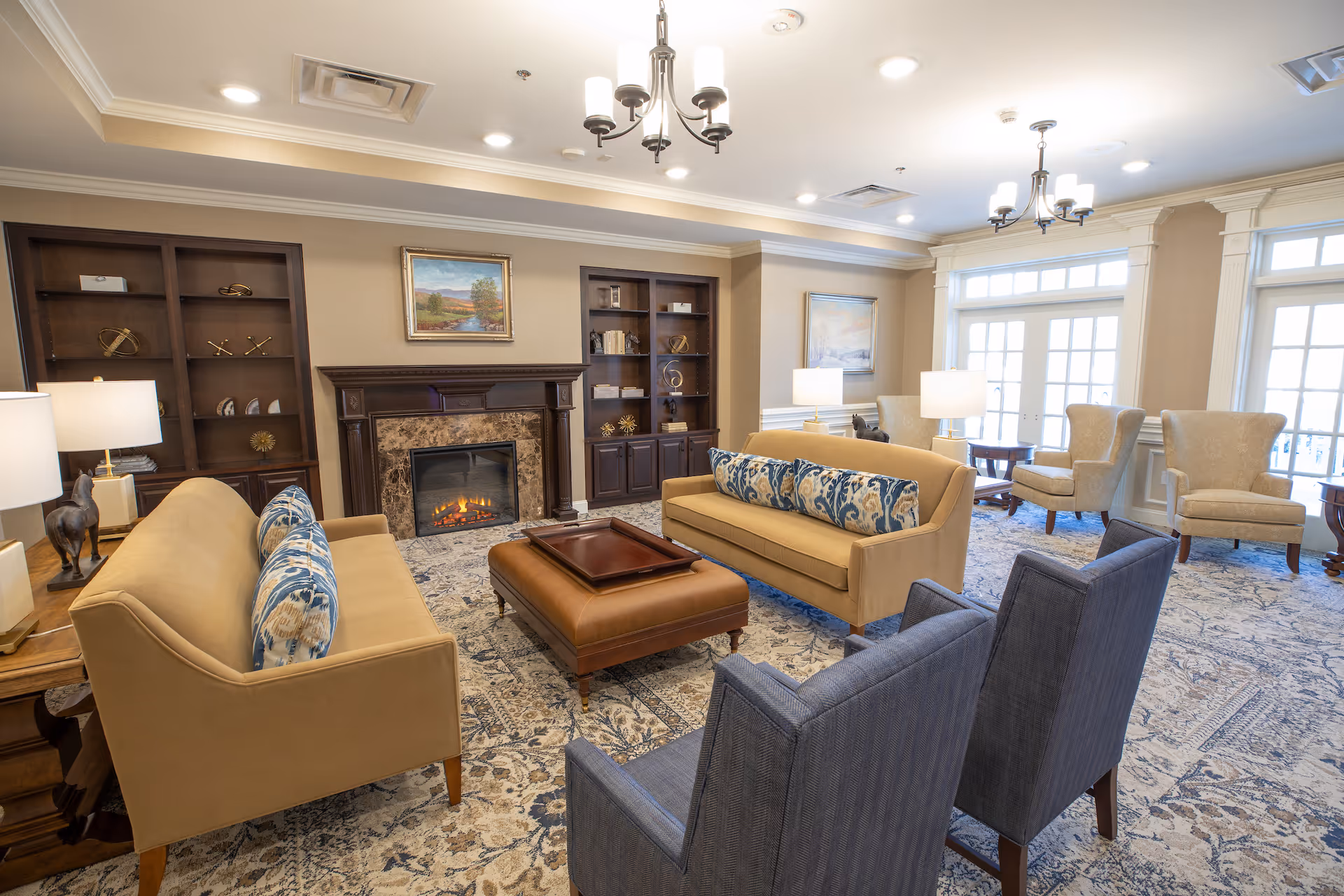 A spacious and well-lit living room featuring two beige sofas with patterned blue and white cushions, two blue armchairs, and two beige wingback chairs near large windows. The room has a decorative fireplace with a painting above it, built-in dark wood bookshelves on either side, a large patterned area rug, and multiple table lamps providing warm lighting. Two chandeliers hang from the ceiling.