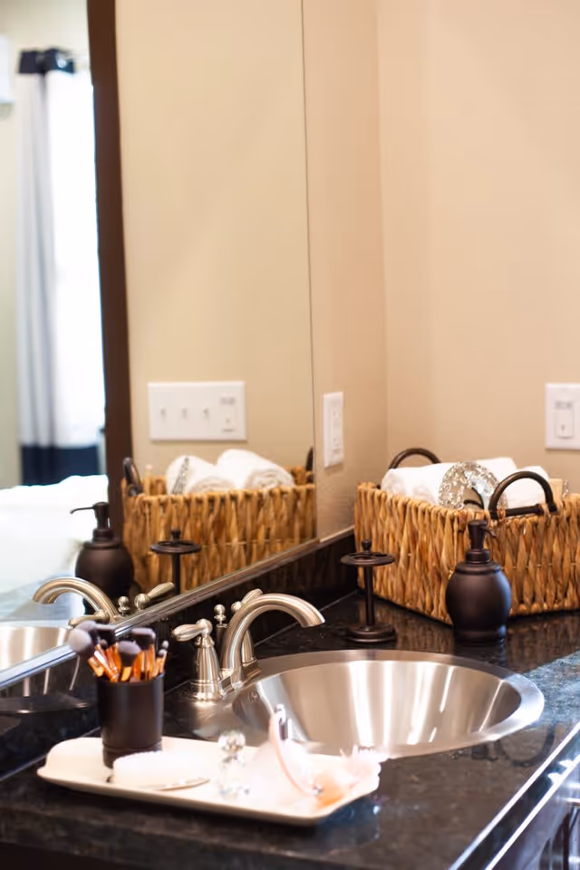 Close-up view of a bathroom sink with a silver faucet on a black countertop. A large mirror reflects the sink area. On the counter, there is a black container holding makeup brushes, a soap dispenser, and a woven basket with rolled white towels and other items.