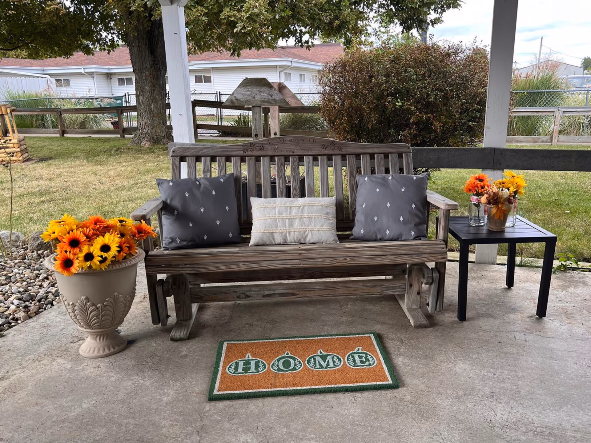 A wooden bench with three cushions placed on a concrete patio under a covered area. To the left of the bench is a large decorative pot filled with bright orange and yellow flowers. To the right is a small black table holding three glass jars with similar orange and yellow flowers. A doormat with the word 'HOME' is placed in front of the bench. In the background, there is a grassy yard with a tree, bushes, and a wooden fence.
