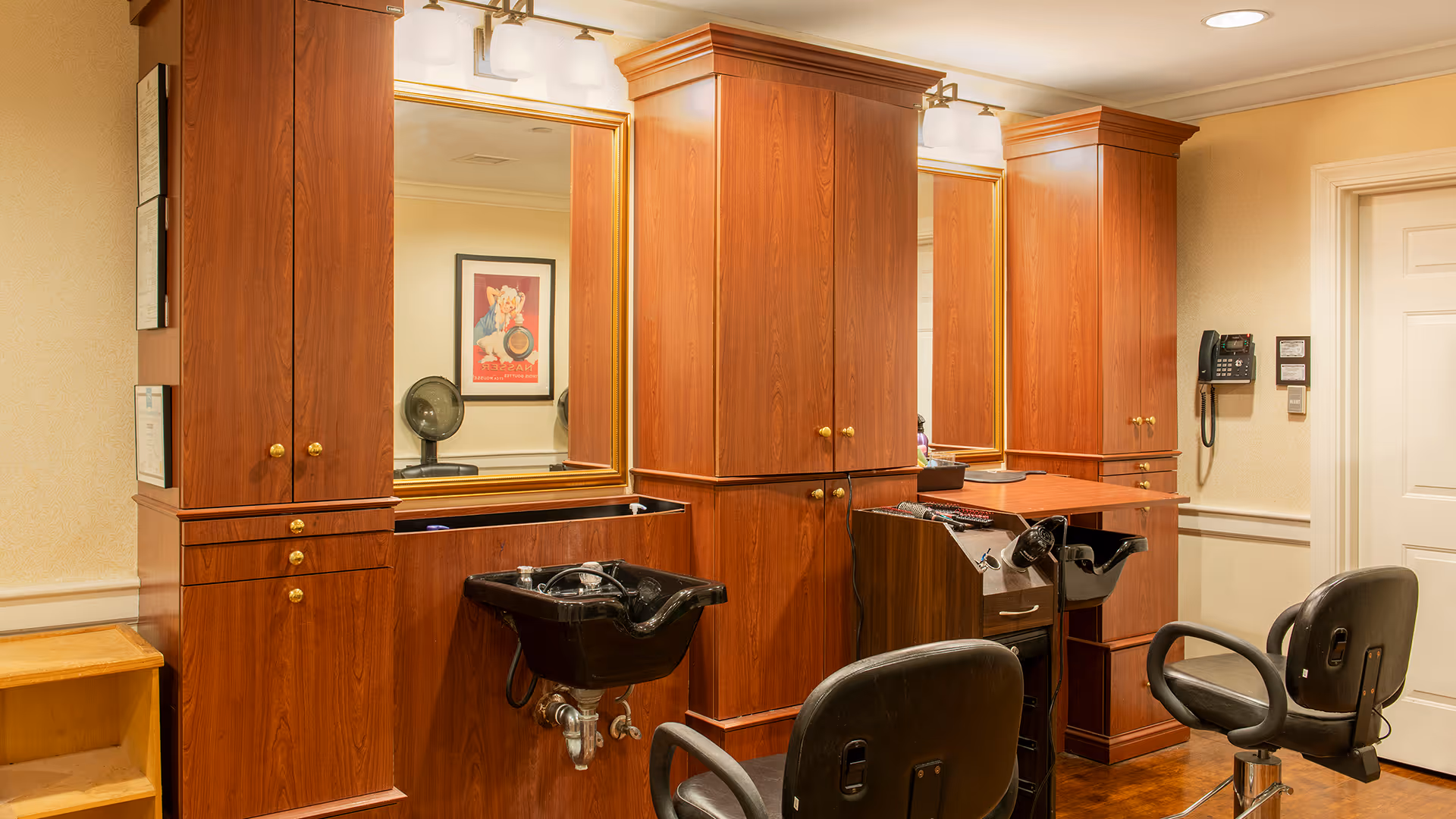 Interior of a salon area with two black salon chairs facing wooden cabinets with mirrors. There are two black wash basins attached to the cabinets for hair washing. The room has beige walls, a framed picture on the wall, and a wall-mounted telephone near a white door.