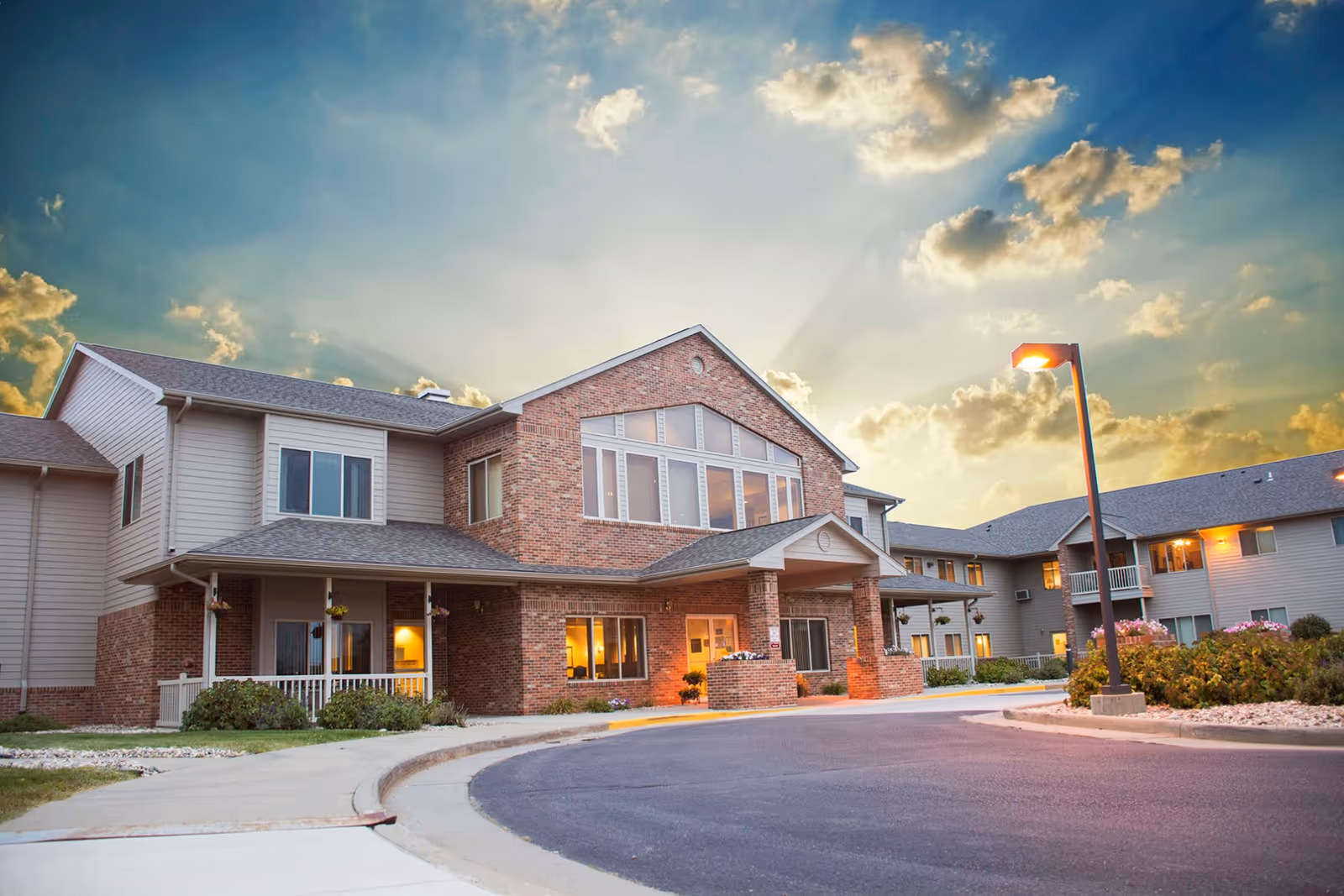 Exterior view of Ponderosa Lodge Senior Living building at sunset with a partly cloudy sky, showing a two-story structure with brick and siding, large windows, a covered entrance, and a curved driveway with street lamps and landscaping.