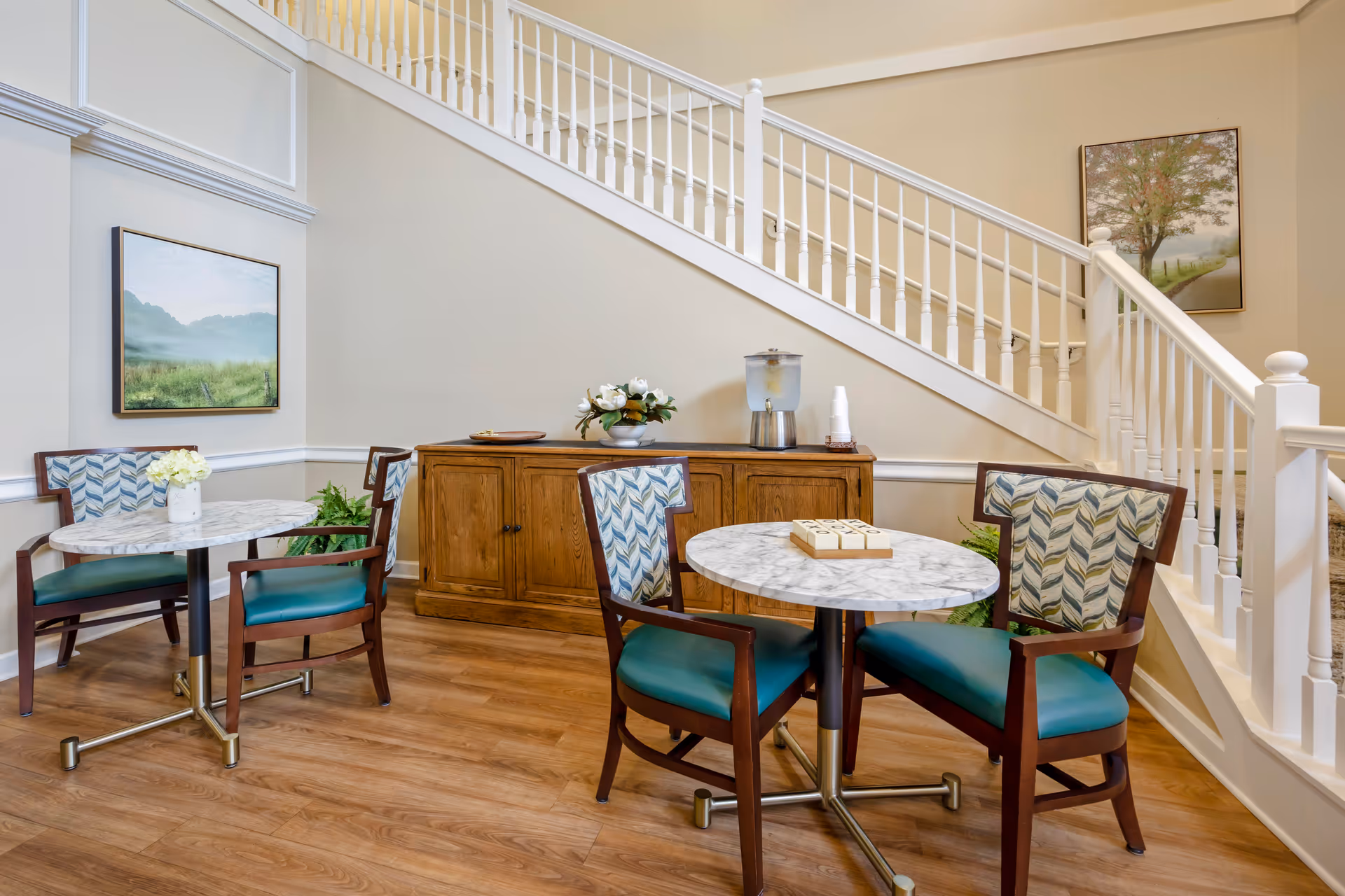 A cozy seating area with two round marble-top tables, each surrounded by two wooden chairs with teal cushions and patterned backs. Behind the tables is a wooden sideboard with a water dispenser, cups, and a flower arrangement. The area is adjacent to a white staircase with wooden handrails and decorated with framed landscape paintings on the walls.