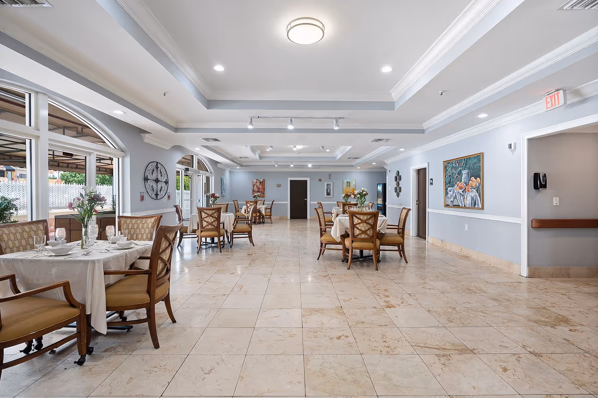 A spacious dining room with several round tables covered with white tablecloths, each set with plates, glasses, and floral centerpieces. The room has large windows on the left side letting in natural light, light blue walls, tiled floors, and ceiling lights. There are paintings and decorative items on the walls, and an exit sign is visible above a doorway on the right.
