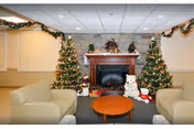 A cozy living room decorated for Christmas with two Christmas trees adorned with lights and ornaments on either side of a fireplace. The fireplace mantel is decorated with holiday greenery and figurines. In front of the fireplace are holiday decorations including a teddy bear and other festive items. The room has beige walls, a drop ceiling with fluorescent lights, and two beige armchairs facing a round wooden coffee table.