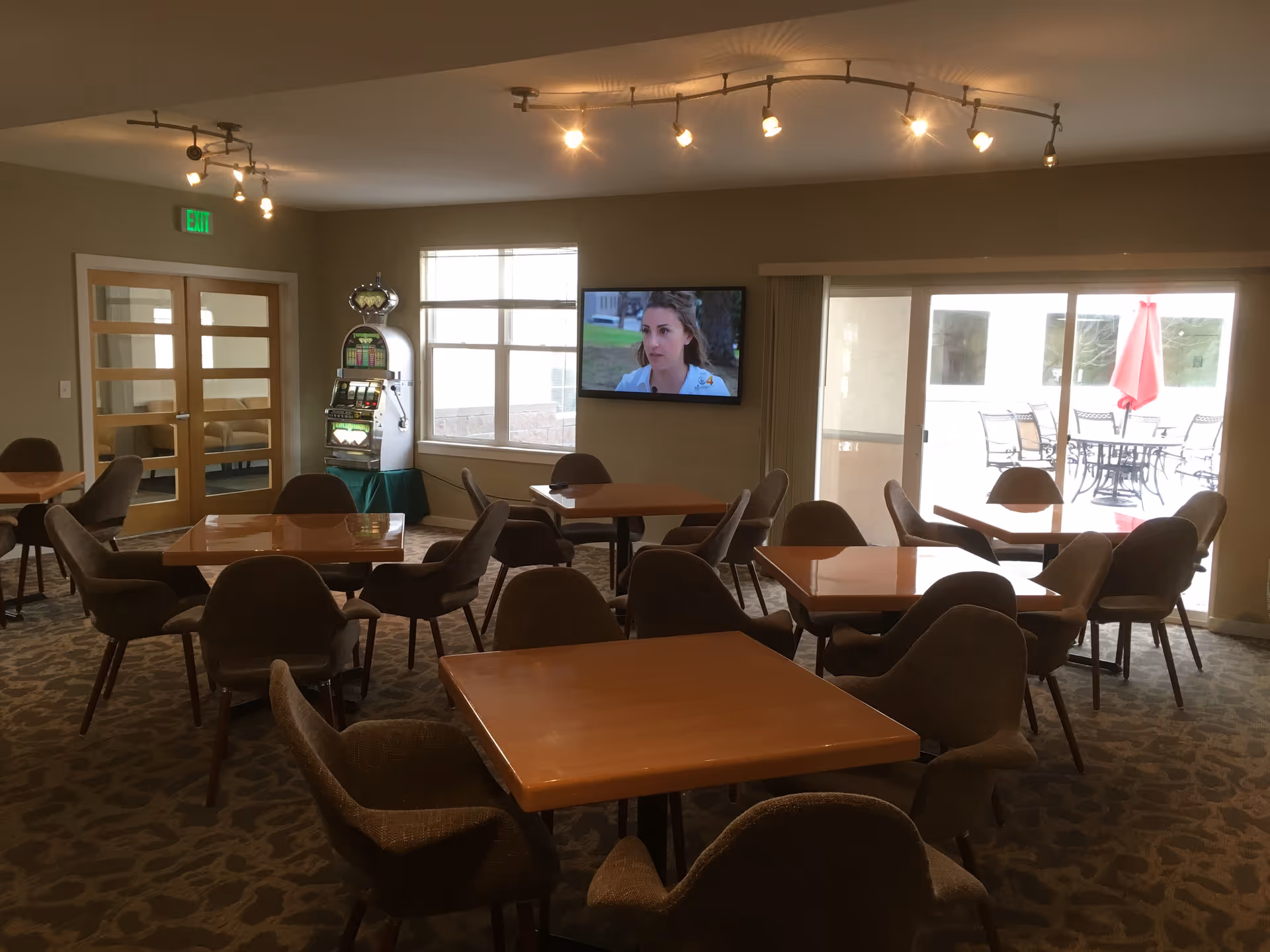 Interior view of a common area with multiple square wooden tables and cushioned chairs arranged around them. A television mounted on the wall displays a woman speaking. There is a slot machine near a window, and glass doors lead to an outdoor patio with tables, chairs, and a red umbrella. The room is lit by ceiling track lights.