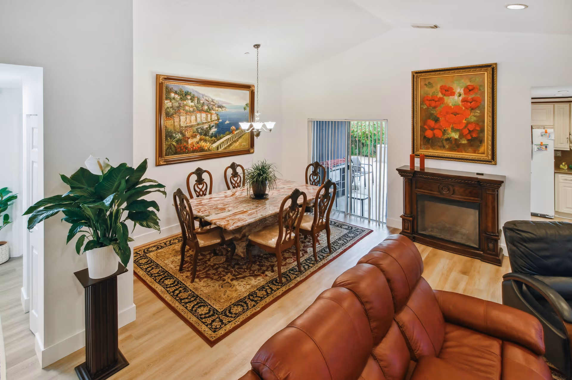 Bright interior showing a dining area with a marble table and six chairs on a decorative rug, adjacent to a leather sofa, fireplace, and wall art.