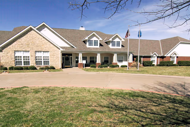 Front exterior view of a single-story senior living facility building with a combination of stone and brick facade, multiple windows, a covered entrance with columns, and two flagpoles displaying the American flag and another flag. The building is surrounded by a lawn and a paved driveway.