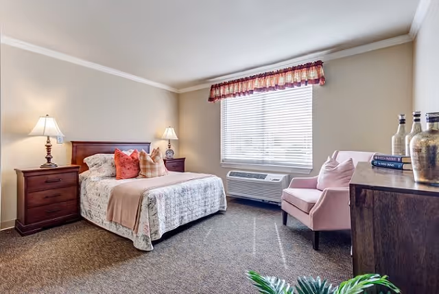 A cozy bedroom in an assisted living facility featuring a bed with floral bedding and multiple pillows, two wooden nightstands with lamps, a pink upholstered armchair, a wooden dresser with decorative vases and books, a large window with blinds and a valance, and a carpeted floor.