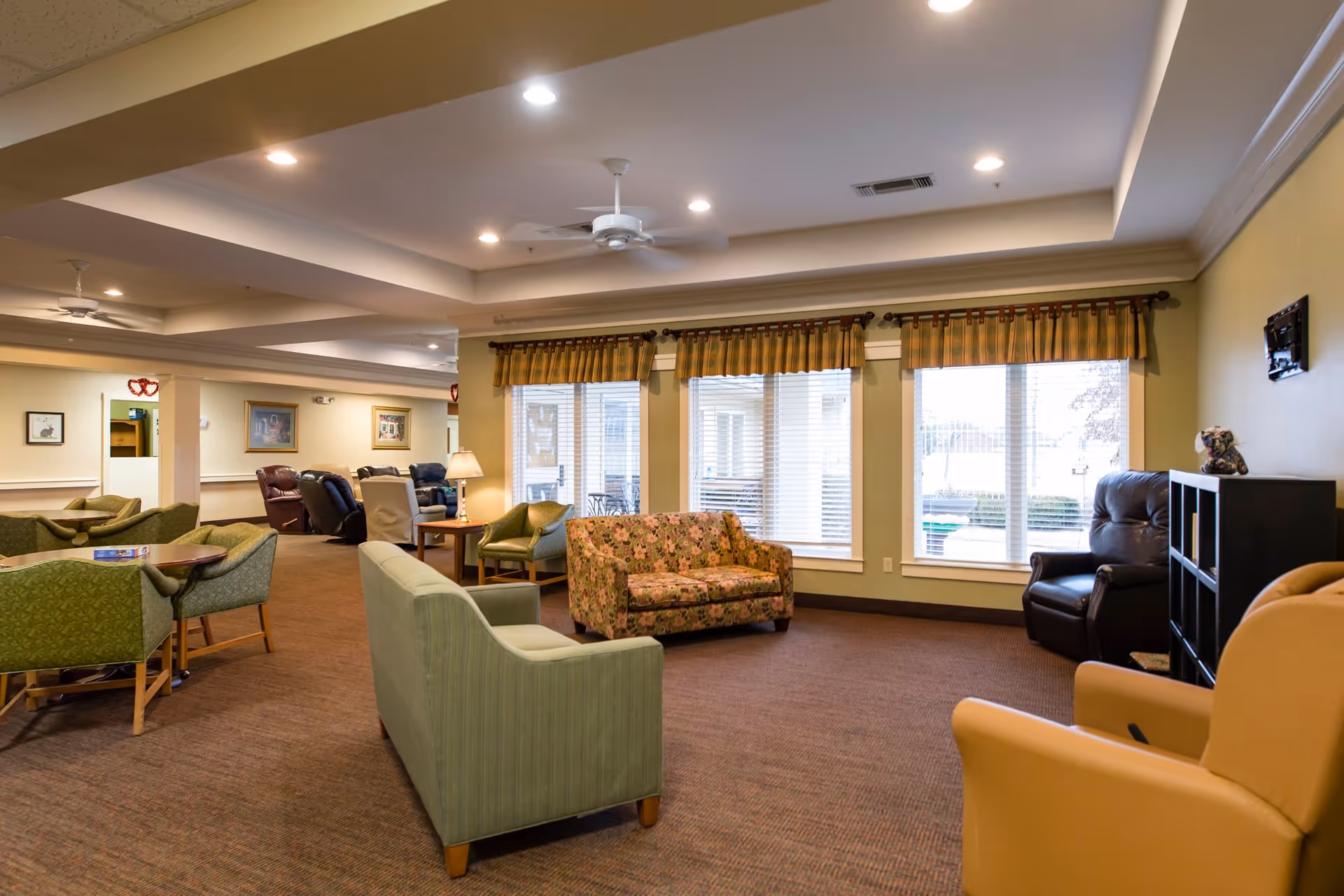 A spacious and well-lit living room area in an assisted living facility featuring multiple seating options including green and floral-patterned sofas, armchairs, and recliners. Large windows with blinds and valances allow natural light to fill the room. The ceiling has recessed lighting and ceiling fans, and the walls are decorated with framed pictures.