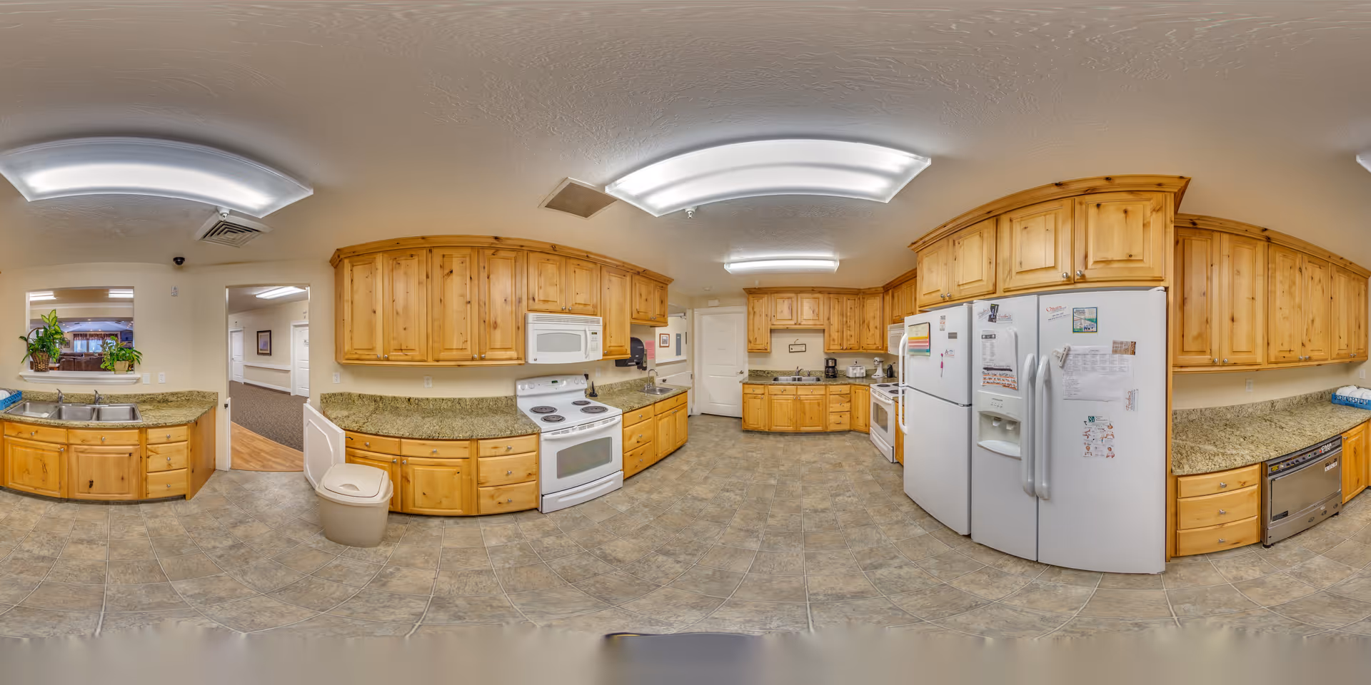 A spacious kitchen with wooden cabinets, granite countertops, and tiled flooring. The kitchen features two white refrigerators, a white stove with an oven, a microwave, a dishwasher, and a double sink. There are fluorescent ceiling lights and a trash can near the sink. The kitchen opens into a hallway and another room with carpeted flooring and plants on the windowsill.