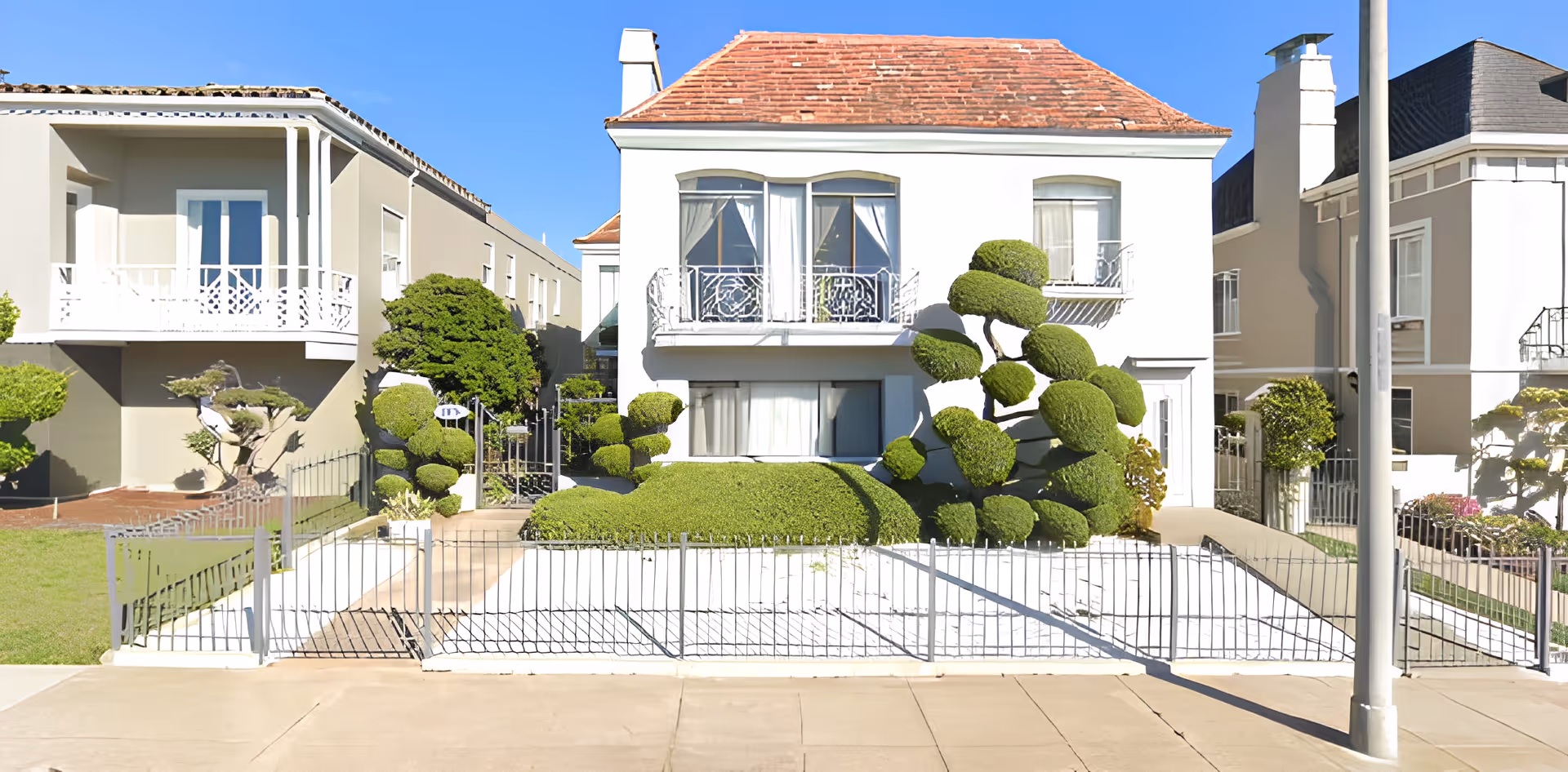 Front exterior view of a white two-story residential building with a red-tiled roof, decorative wrought iron balcony railings, and neatly trimmed topiary bushes in the front yard, surrounded by a low metal fence.