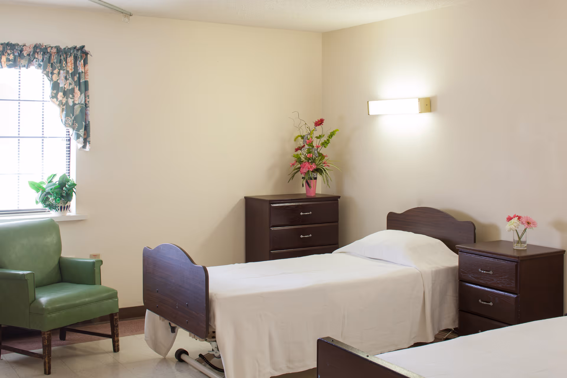 A senior living bedroom with a single bed, wooden nightstands and dresser, a green armchair by a window, and floral arrangements.