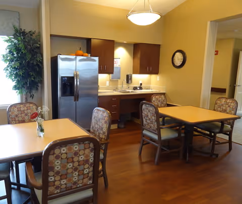 A small dining area with two square tables and patterned chairs. In the background, there is a kitchenette with a stainless steel refrigerator, wooden cabinets, a sink, and a clock on the wall. A potted plant is visible near a window with curtains.